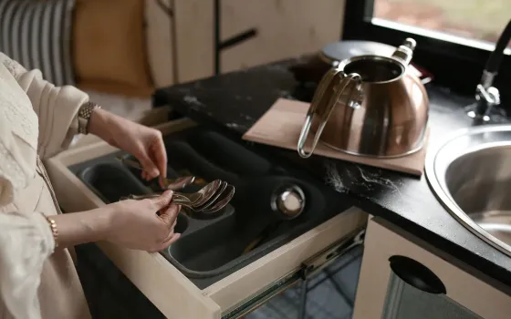 Woman lifting spoons from a grey cutlery tray in an open drawer beside a steel hob-top kettle