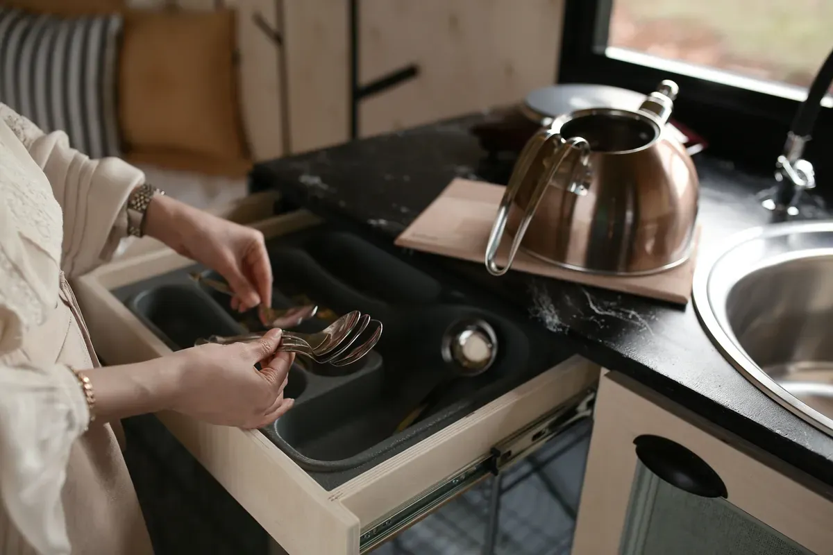 Woman lifting spoons from a grey cutlery tray in an open drawer beside a steel hob-top kettle