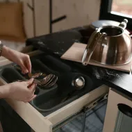 Woman lifting spoons from a grey cutlery tray in an open drawer beside a steel hob-top kettle