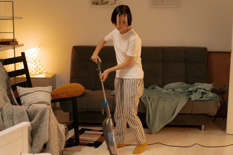 Person in striped pyjama trousers running a stick vacuum over a rug beside a dark grey sofa
