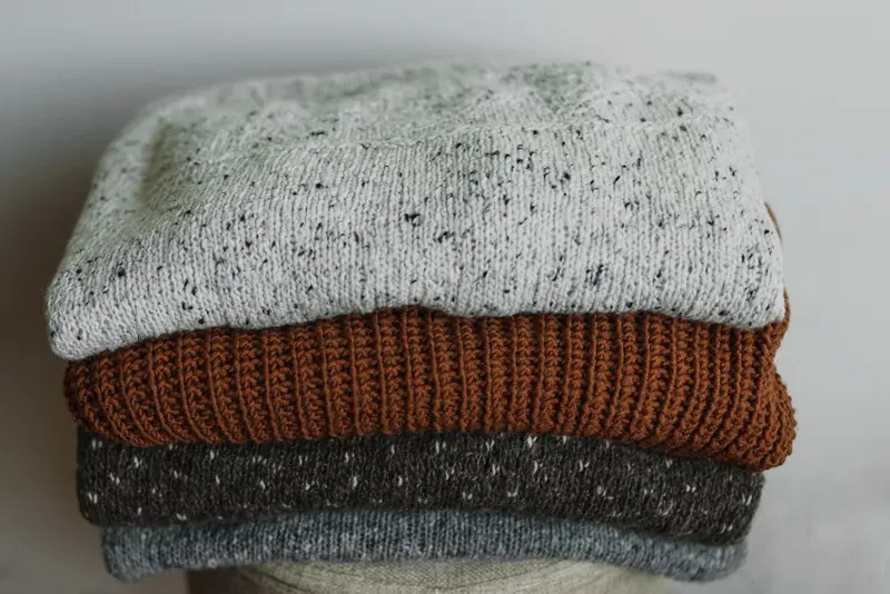 Stack of four folded jumpers, flecked grey, rust-brown ribbed and charcoal, on a pale surface