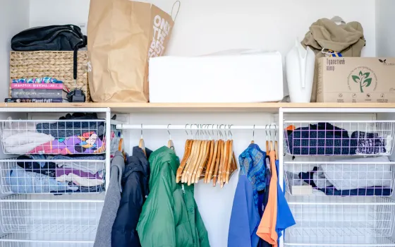 Open wardrobe with a green puffer jacket on wooden hangers, wire basket drawers and a paper bag above