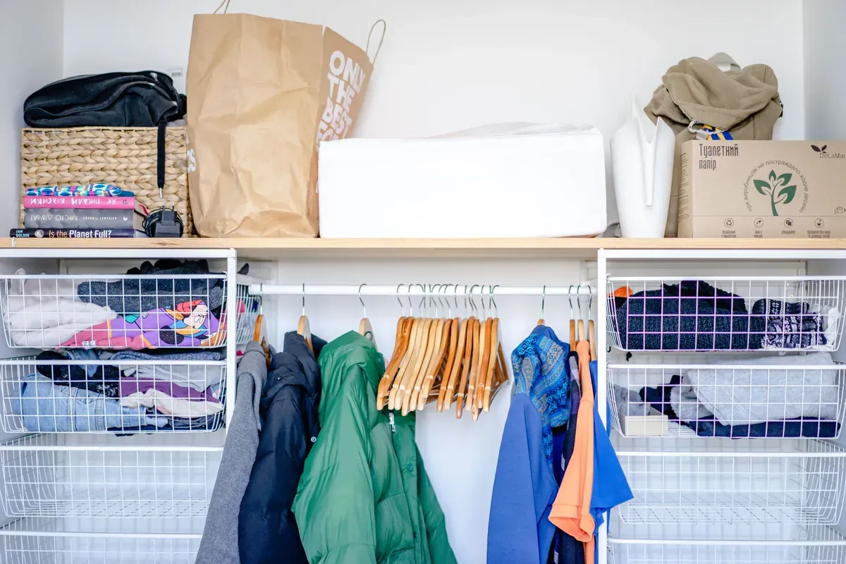 Open wardrobe with a green puffer jacket on wooden hangers, wire basket drawers and a paper bag above