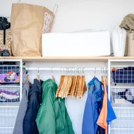 Open wardrobe with a green puffer jacket on wooden hangers, wire basket drawers and a paper bag above