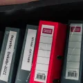 Row of grey lever-arch files with a single red one, spines labelled by month, on a dark shelf