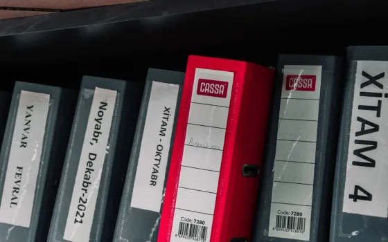 Row of grey lever-arch files with a single red one, spines labelled by month, on a dark shelf