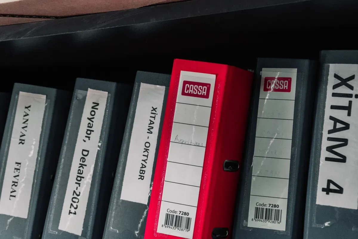 Row of grey lever-arch files with a single red one, spines labelled by month, on a dark shelf