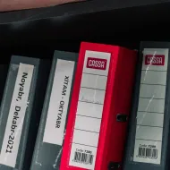 Row of grey lever-arch files with a single red one, spines labelled by month, on a dark shelf