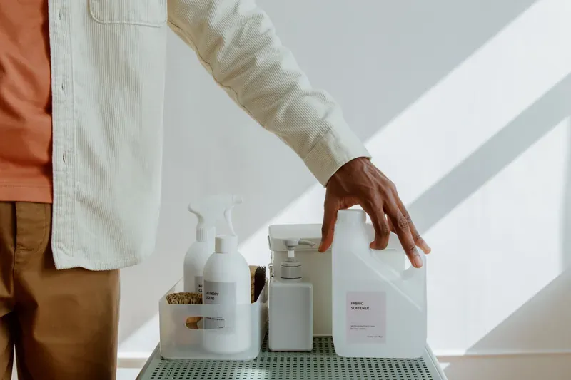 Man in a cream corduroy shirt reaching for a white bottle of fabric softener on a tray of cleaning products