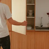 Man in a white T-shirt opening a pale wooden wall cupboard of cups and plates above a steel sink