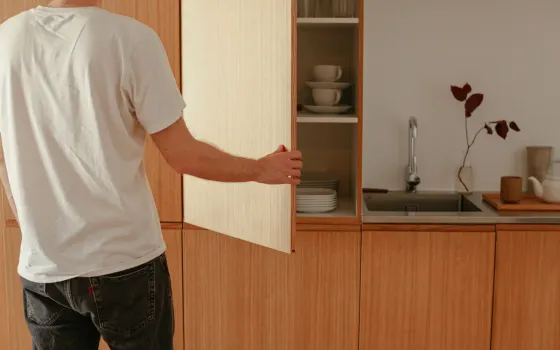 Man in a white T-shirt opening a pale wooden wall cupboard of cups and plates above a steel sink