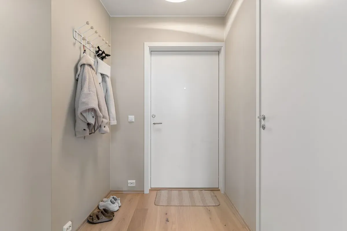 Narrow beige hallway with a white front door, two fleece jackets on wall hooks and trainers on the floor
