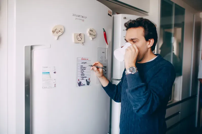 Man in a navy cable-knit jumper drinking from a white mug while writing on a notepad stuck to a white fridge door