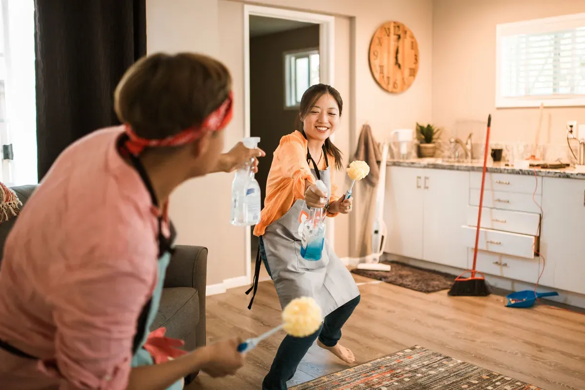 Two people in grey aprons play-fighting with a spray bottle and yellow dusters in a kitchen with a wooden wall clock