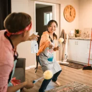 Two people in grey aprons play-fighting with a spray bottle and yellow dusters in a kitchen with a wooden wall clock