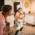 Two people in grey aprons play-fighting with a spray bottle and yellow dusters in a kitchen with a wooden wall clock
