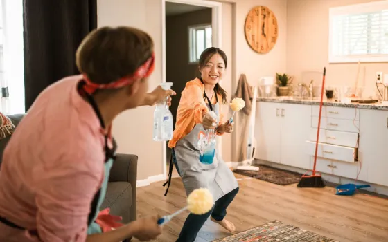 Two people in grey aprons play-fighting with a spray bottle and yellow dusters in a kitchen with a wooden wall clock