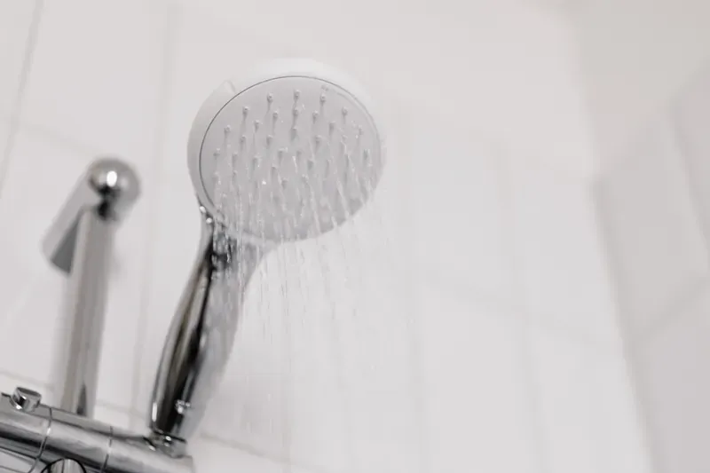 Chrome hand-held shower head spraying water against white square wall tiles, with a mixer bar below