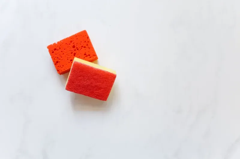 Two orange kitchen sponges, one with a red scouring pad on top, on a white marble surface