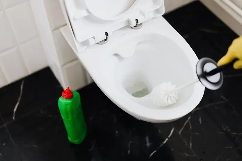 Person in yellow rubber gloves scrubbing a white toilet with a brush, a green cleaner bottle on a black marble floor