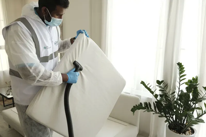 Man in a white protective suit and blue gloves running a vacuum nozzle over an upended white mattress