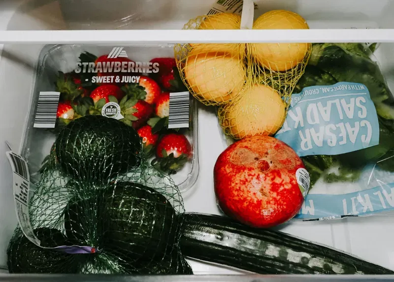Fridge salad drawer holding a punnet of strawberries, netted lemons and avocados, a pomegranate, a cucumber and a bagged salad kit