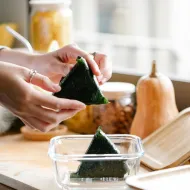 Hands placing a nori-wrapped rice triangle into a glass container beside a squash and jars