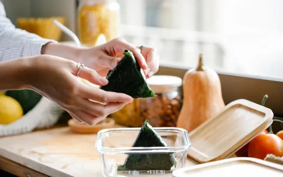 Hands placing a nori-wrapped rice triangle into a glass container beside a squash and jars