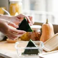 Hands placing a nori-wrapped rice triangle into a glass container beside a squash and jars