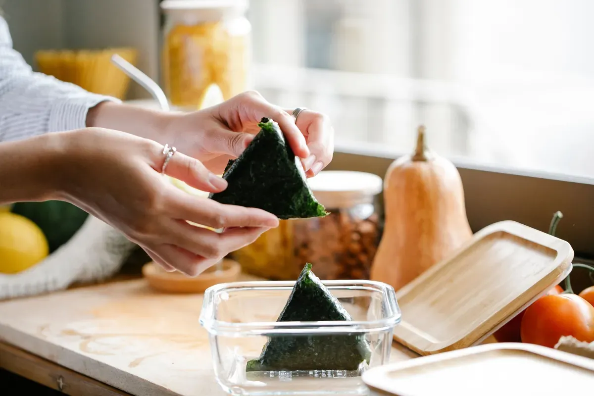 Hands placing a nori-wrapped rice triangle into a glass container beside a squash and jars