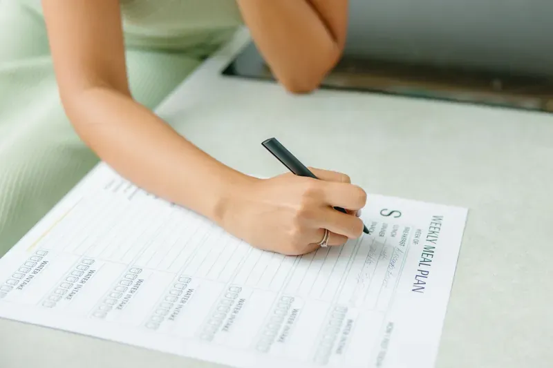 Woman in a pale green top writing on a printed weekly meal plan sheet with a black pen