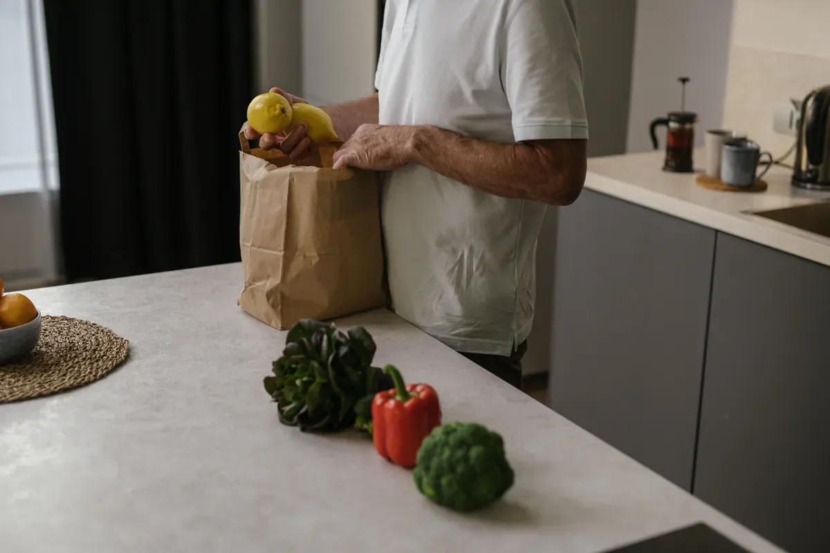 Man in a white T-shirt lifting lemons from a paper bag, with broccoli and a red pepper on the worktop