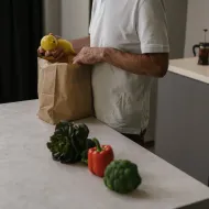 Man in a white T-shirt lifting lemons from a paper bag, with broccoli and a red pepper on the worktop