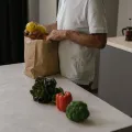 Man in a white T-shirt lifting lemons from a paper bag, with broccoli and a red pepper on the worktop