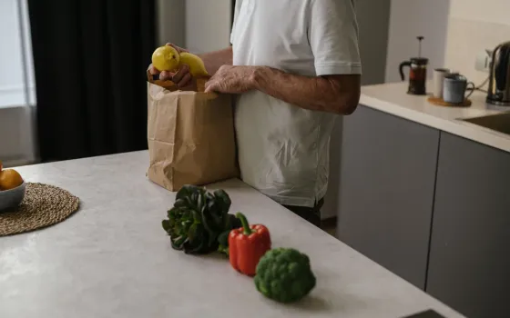 Man in a white T-shirt lifting lemons from a paper bag, with broccoli and a red pepper on the worktop