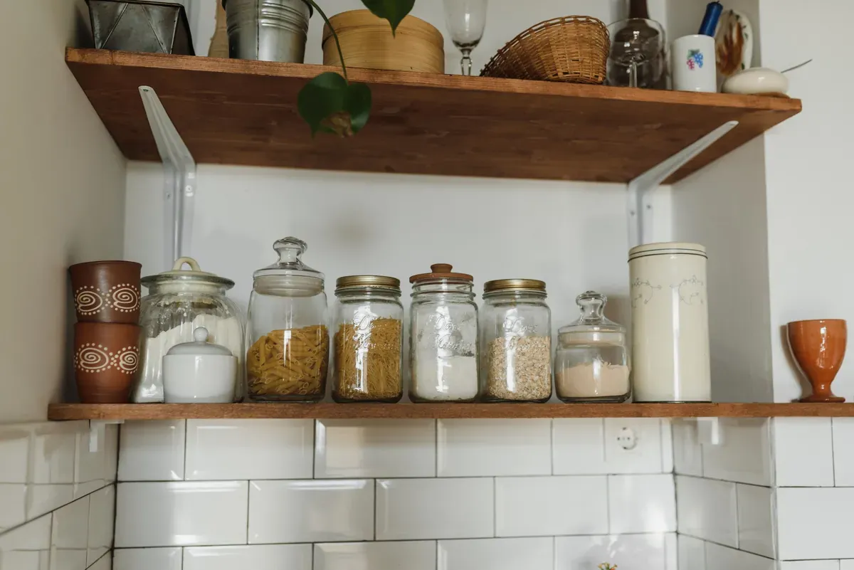 Glass jars of pasta, spaghetti, flour and oats on a wooden shelf above white metro tiles