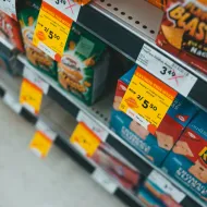 Angled view of yellow Savings This Week stickers clipped to a shelf edge under boxes of snack crackers