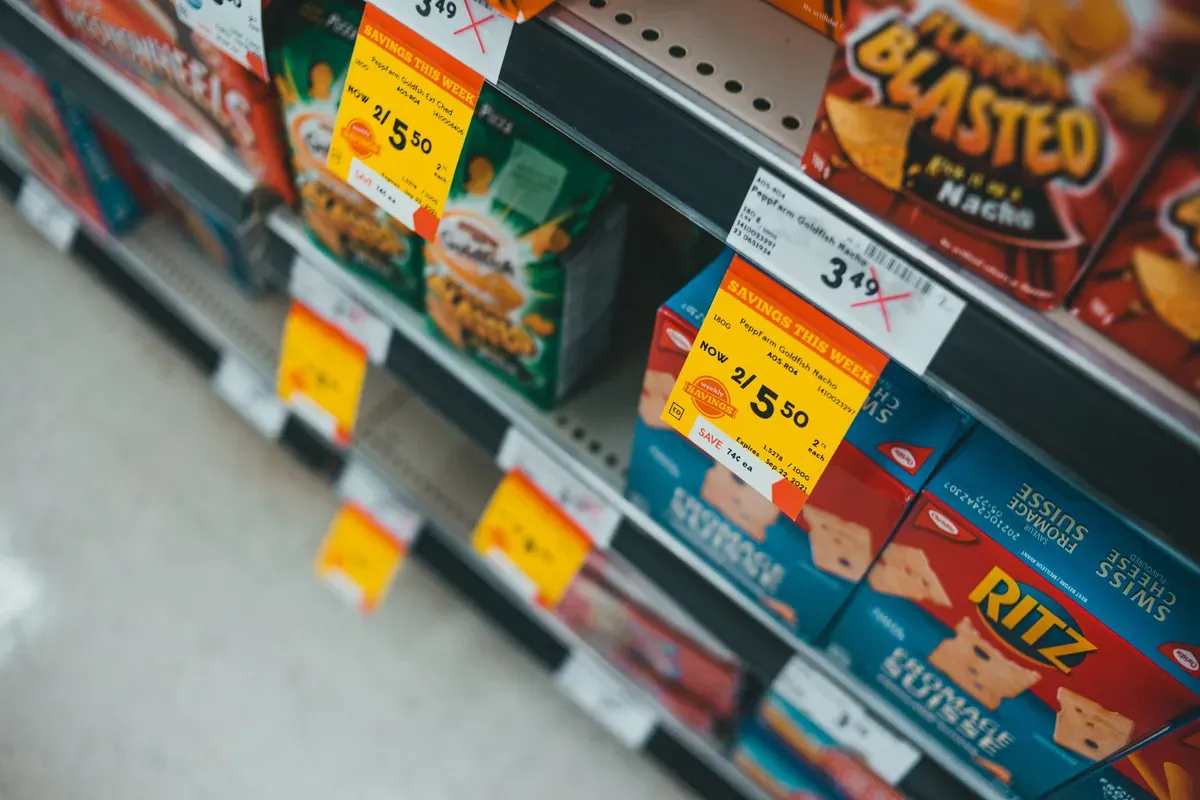 Angled view of yellow Savings This Week stickers clipped to a shelf edge under boxes of snack crackers