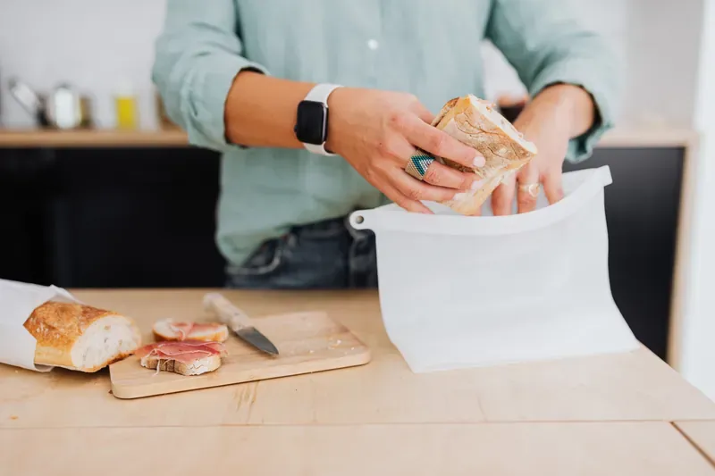 Person in a green shirt putting a half baguette into a white reusable food bag, with a knife and ham on a wooden board