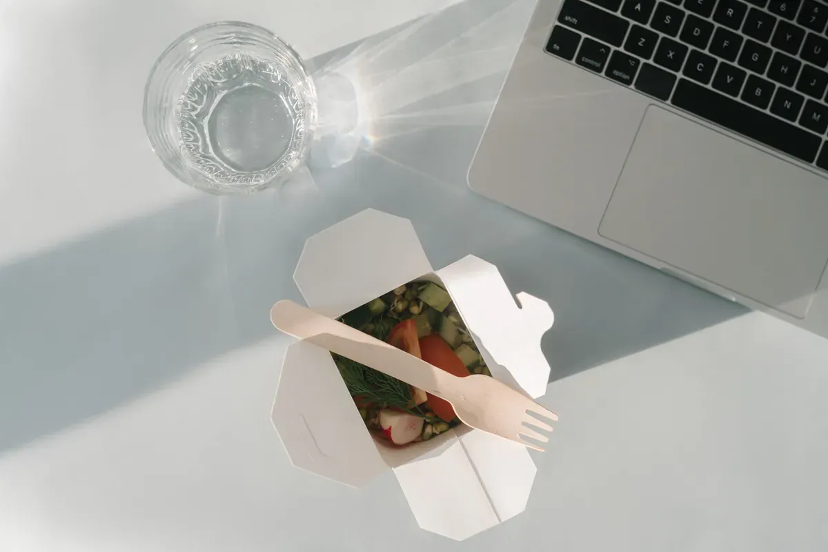 Open white takeaway carton of chopped salad with a wooden fork, next to a laptop keyboard and a glass of water