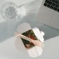 Open white takeaway carton of chopped salad with a wooden fork, next to a laptop keyboard and a glass of water