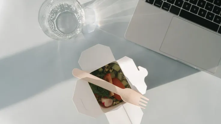 Open white takeaway carton of chopped salad with a wooden fork, next to a laptop keyboard and a glass of water
