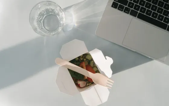 Open white takeaway carton of chopped salad with a wooden fork, next to a laptop keyboard and a glass of water