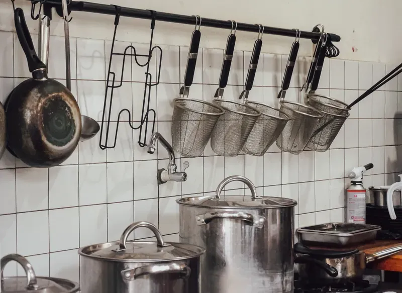 Black rail with S-hooks holding a wok and five wire strainers above stainless steel stockpots on a tiled wall