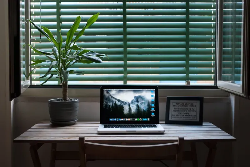 Slatted wooden desk under a window with green venetian blinds, holding a laptop, a potted plant and a framed sign
