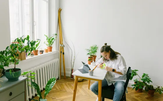 Woman writing at a small white table beside a radiator and window sill lined with potted plants