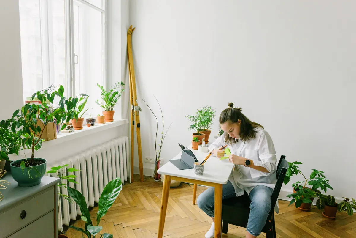 Woman writing at a small white table beside a radiator and window sill lined with potted plants
