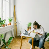 Woman writing at a small white table beside a radiator and window sill lined with potted plants