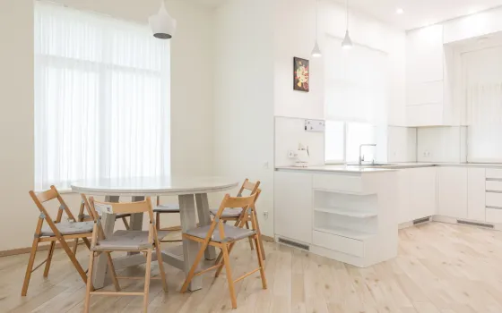 Round white dining table with four wooden folding chairs in a white kitchen with sheer curtains