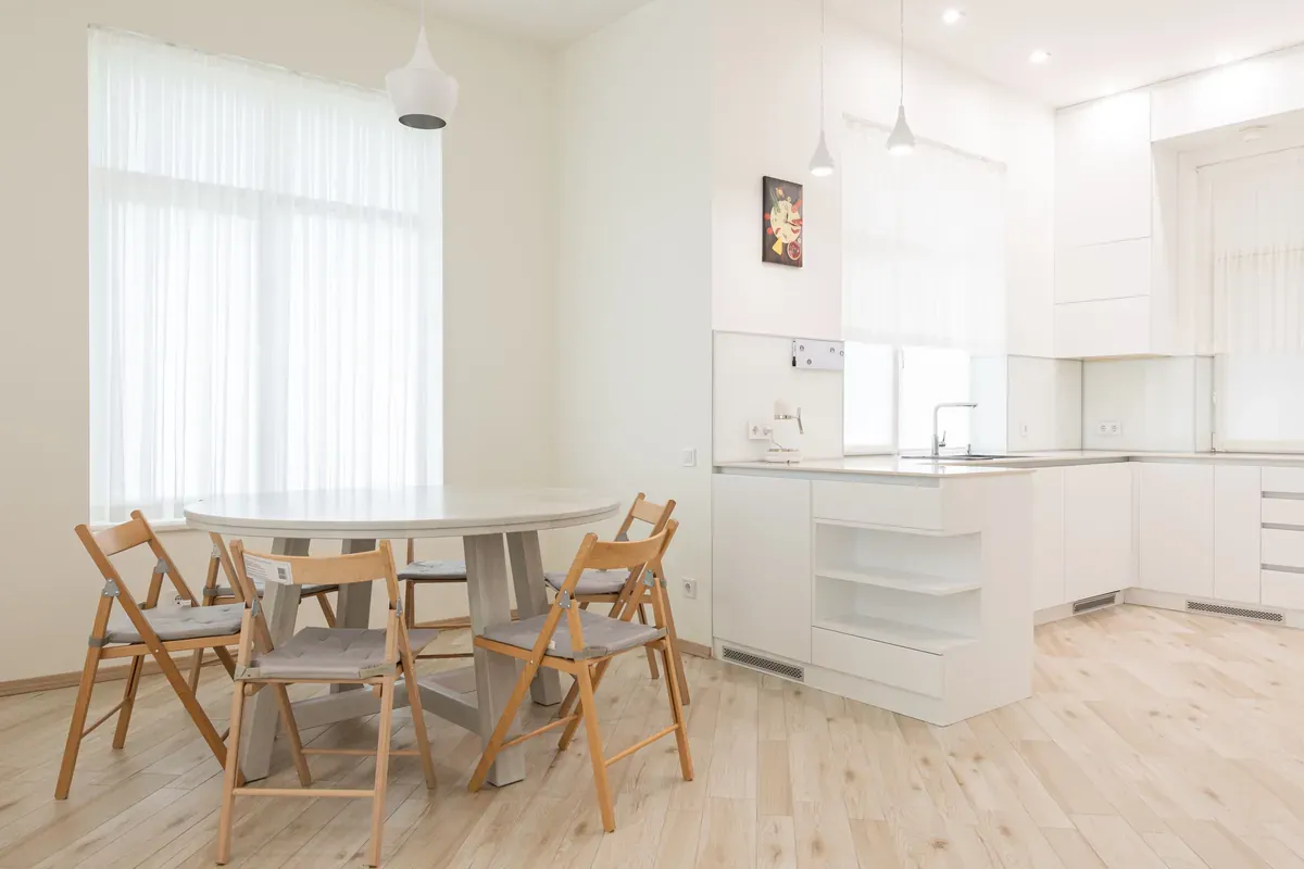 Round white dining table with four wooden folding chairs in a white kitchen with sheer curtains